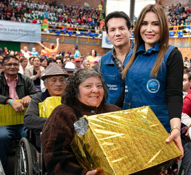 El Dr. César Moreno y la Dra. Ángela Oviedo entregando un paquete dorado a una persona con discapacidad en un evento, rodeados de una multitud en un coliseo.